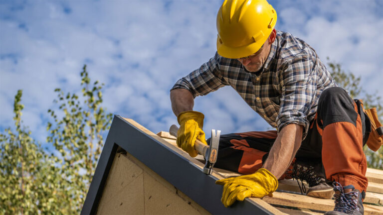 A man in yellow gloves and hard hat uses a hammer while working on a roof.