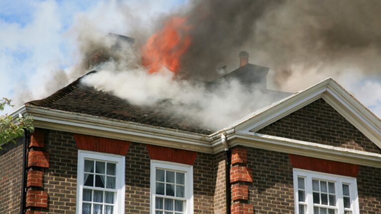A roof with shingles is on fire with dark smoke around it.