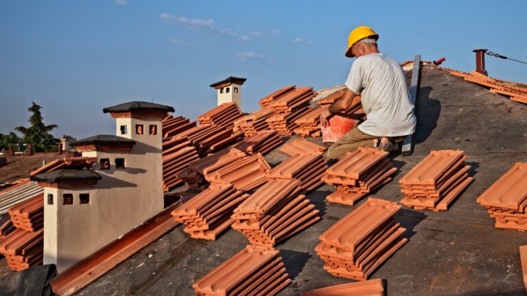 A roofing contractor sits on a roof surrounded by piles of tiles.